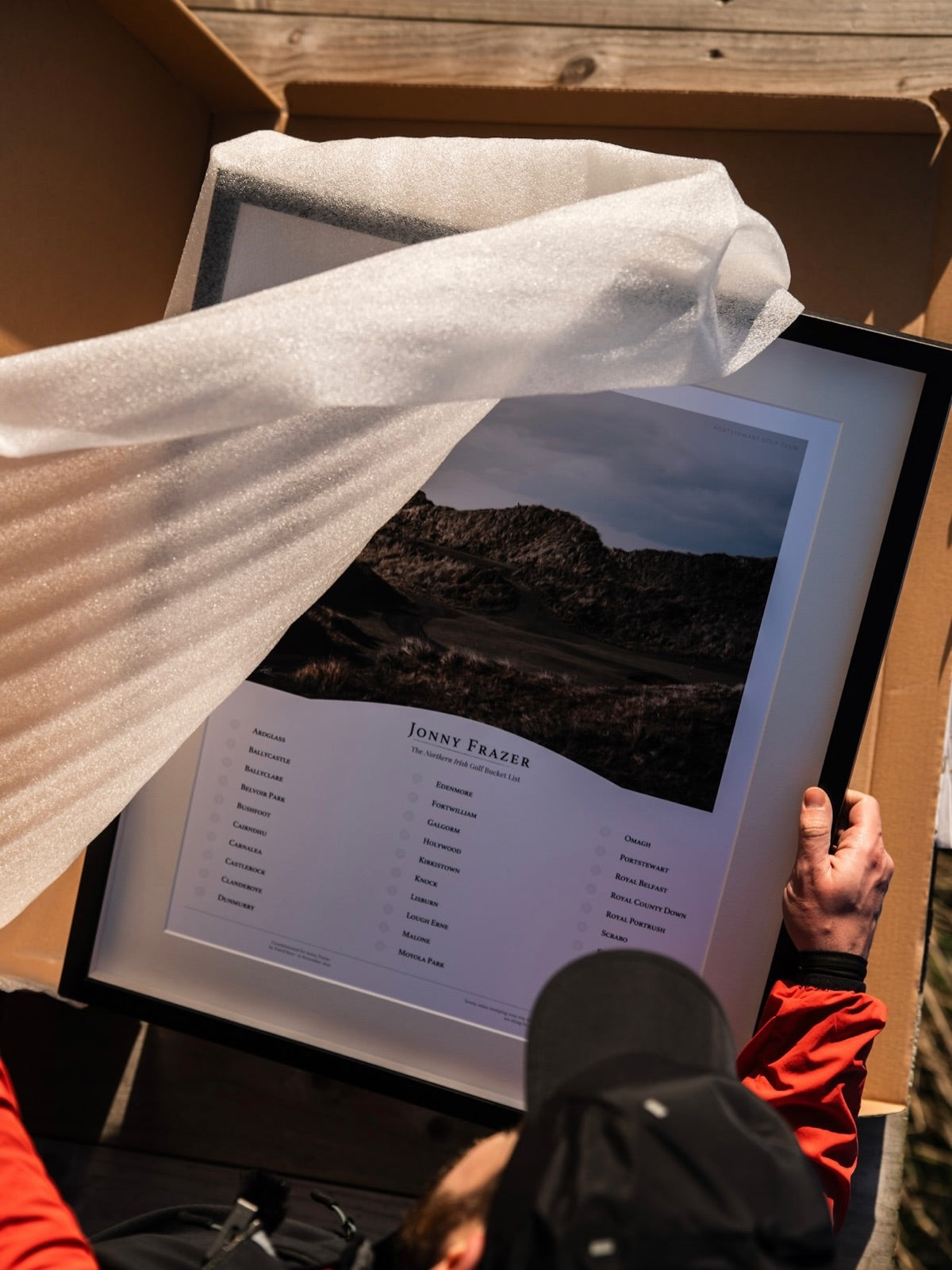 A framed BHÍOS bucket list piece still in its box, bubble wrap pulled back to show the print and its black timber frame.
